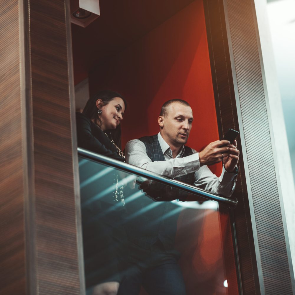 A handsome caucasian businessman is leaning on a chrome handrail of an office balcony and showing the screen of his smartphone to his female business colleague while they both having a conversation